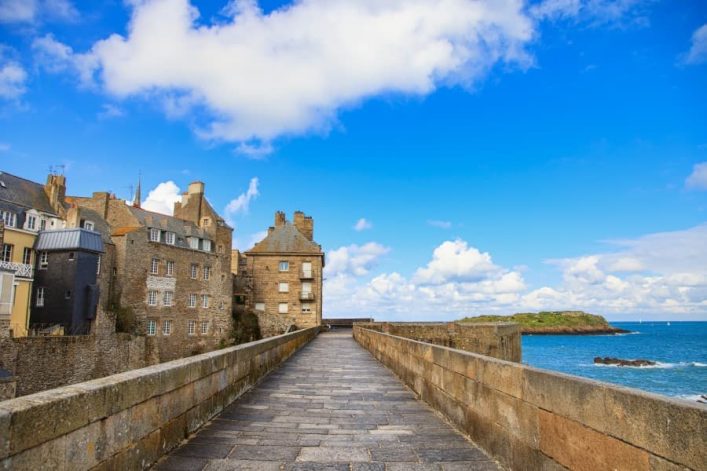 Foto aufgenommen auf der Stadtmauer in Saint Malo. Rechts ist das Meer zu sehen, links liegt die Stadt. Der Himmel ist blau mit einigen Wolken durchzogen.