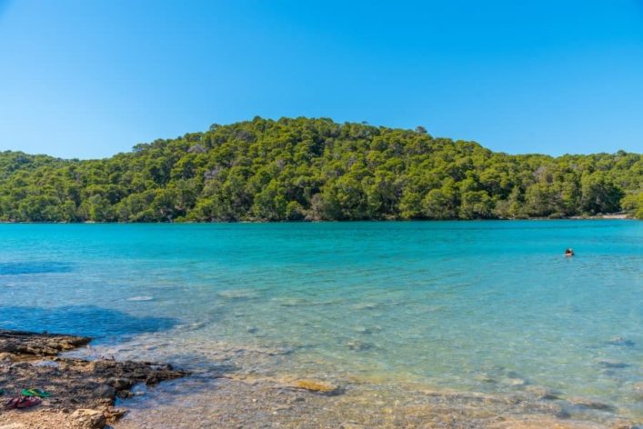 Auf dem Bild sieht man das türkisblaue Wasser des Malo Jezero Sees. Dahinter sind die grünen Bäume des Nationalparks Mljet zu sehen.
