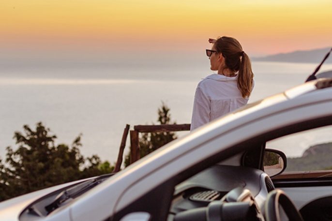 Eine Frau hat mit ihrem Auto vor einer eindrucksvollen Kulisse angehalten und bestaunt den Ausblick auf einen zauberhaften orange-roten Sonnenuntergang, der sich im ruhigen Meer spiegelt. Mit Vordergrund ist das Auto zu sehen, dessen Türen noch offen sind. Es schaut so aus, als hätte die Frau es kaum abwarten können, sich dieses Schauspiel am Horizont anzusehen. Im Hintergrund sieht man den Umriss einer bergigen Landschaft.