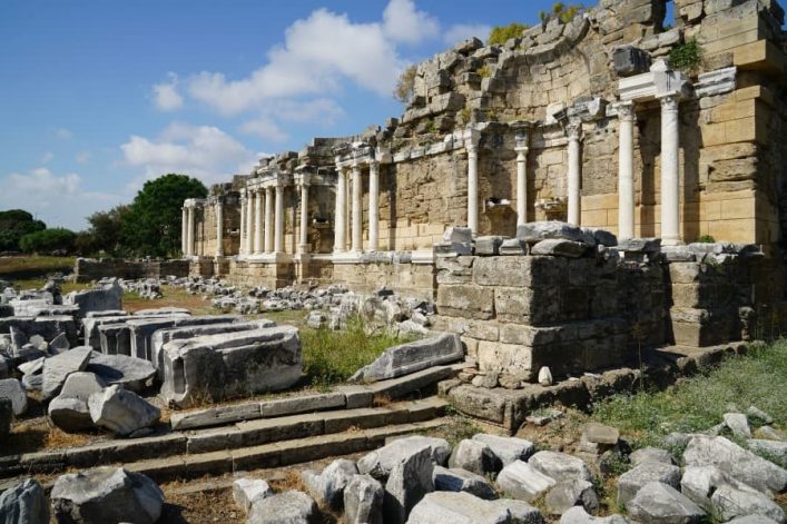 Antike Ruinen in Side mit Säulen und Steinen, blauer Himmel, Grünpflanzen und Bruchstücke im Vordergrund, Agora und Tempelfassade im Hintergrund.