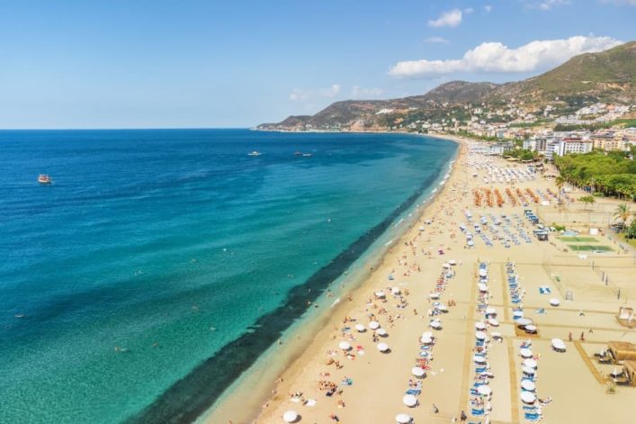 Luftaufnahme des Kleopatra-Strands in Side: Türkisblaues Meer grenzt an goldgelben Sandstrand, gesäumt von Sonnenschirmen und Liegestühlen, mit grünen Hügeln im Hintergrund.