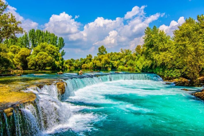 Türkisfarbener Wasserfall im Grünen, umgeben von üppigen Bäumen, strahlt unter blau-weißem Himmel - ein Highlight in Side.
