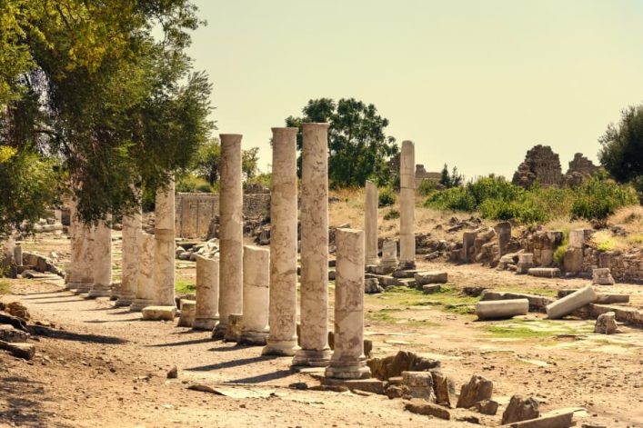 Antike Säulenreihe in Side, Türkei, umgeben von Ruinen und Grün, auf Sandweg unter sonnigem Himmel.