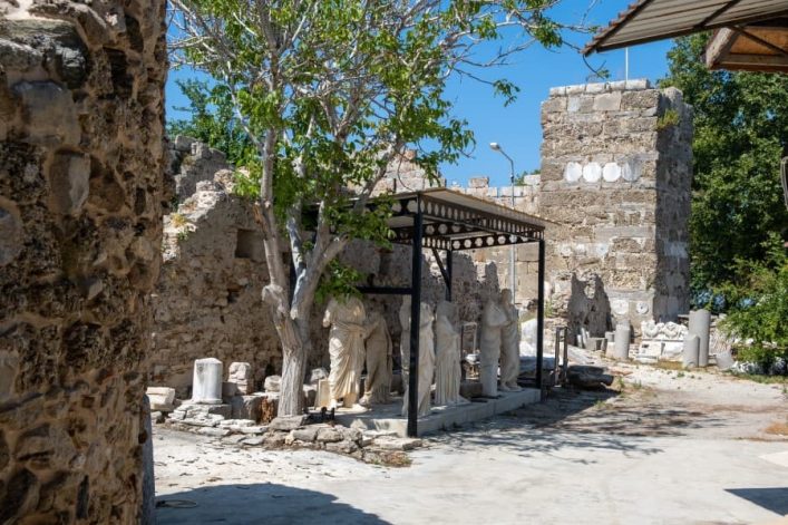 Antike Statuen und Säulenstümpfe unter einem Schutzdach im Side Museum, umgeben von historischen Ruinen und begrüntem Baum im Vordergrund.