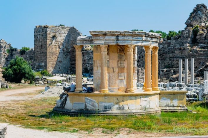 Rundes Monument aus Marmor mit korinthischen Säulen, Tyche-Tempel in Side. Antike Ruinen und grüne Vegetation im Hintergrund unter blauem Himmel.