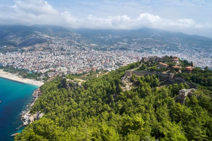 Blick auf Alanya mit der imposanten Burganlage, umgeben von grünen Bäumen, oberhalb der blauen Küstenlinie und der dicht bebauten Stadt.