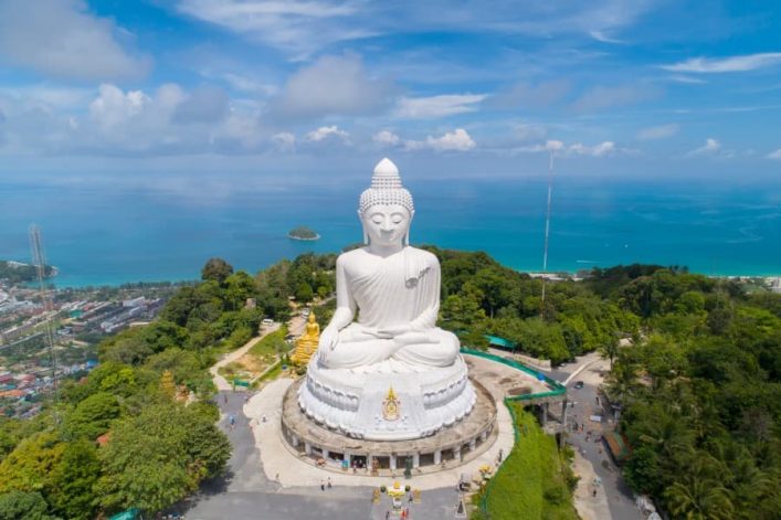 Der gigantische weiße Big Buddha von Phuket thront majestätisch auf einem Hügel mit üppigem Grün im Vordergrund. Im Hintergrund erstreckt sich das türkisblaue Meer als beliebte Kreuzfahrtroute Richtung Thailand.