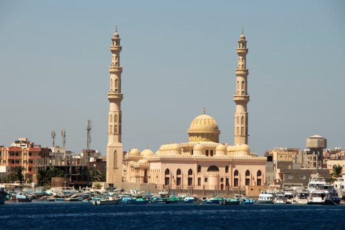 Die Al-Mina-Moschee in Hurghada mit goldener Kuppel und zwei Minaretten, umgeben von Booten auf dem Wasser und Gebäuden im Hintergrund.