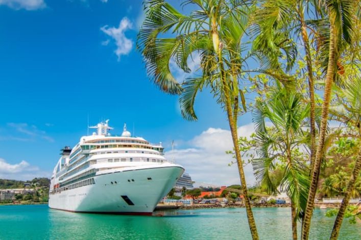 Landschaft mit tropischen Palmen, einem großen weißen Kreuzfahrtschiff im Hafen und einer belebten Küstenlinie mit Bergen im Hintergrund.