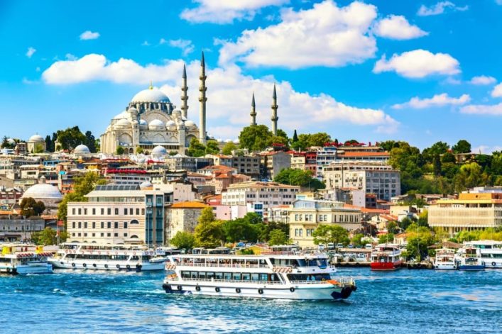Stadtkulisse am Wasser in Istanbul mit der prächtigen Hagia Sophia im Hintergrund auf einem Hügel. Im Hafen, beliebte Kreuzfahrtrouten, ankern und fahren mehrere weiße Boote. Ein strahlend blauer Himmel mit vereinzelten Wolken rundet die Szenerie ab.