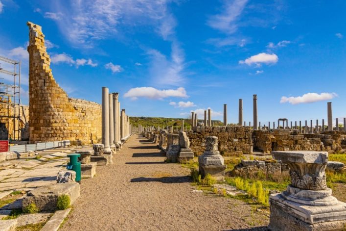 Alte Ruinen von Perge nahe Antalya. Links stehen restaurierte Säulen und die Überreste einer hohen Mauer aus gelben Steinen, rechts weitere Säulen und verfallene Steinstrukturen bei blauem Himmel.