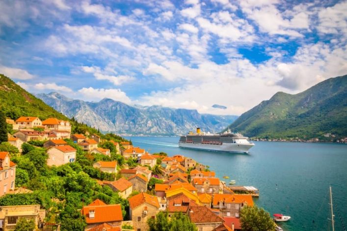 Eine Kreuzfahrt führt vorbei an einer malerischen Küstenstadt mit roten Dächern, grüne Hügel und majestätische Berge im Hintergrund. Ein Kreuzfahrtschiff passiert die Bucht.