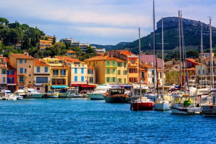 Bunte Häuser mit roten Dächern am Hafen vor bewaldeten Hügeln. Zahlreiche Boote und Jachten im Wasser. Beliebte Kreuzfahrtrouten führen oft zu solch malerischen Küstenorten.