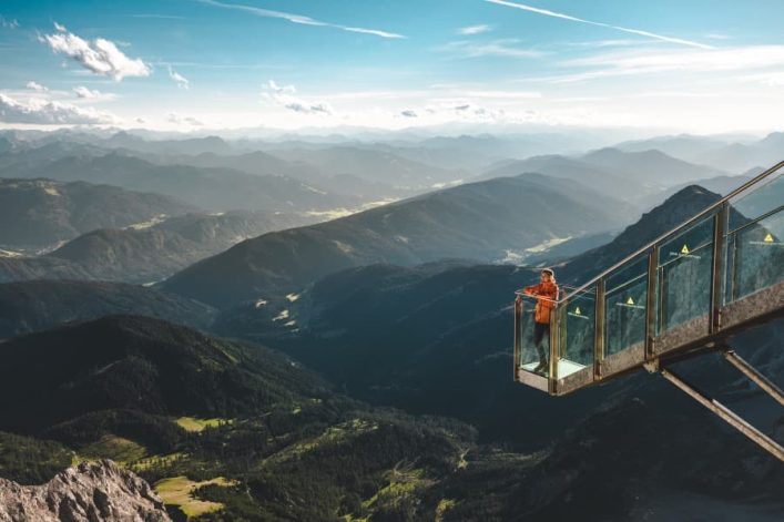 Person auf gläserner Plattform schaut auf ein weites, grünes Tal, umgeben von hohen, bewaldeten Bergen. Im Vordergrund links Felsen, im Hintergrund blauer Himmel mit weißen Wolken und Kondensstreifen.
