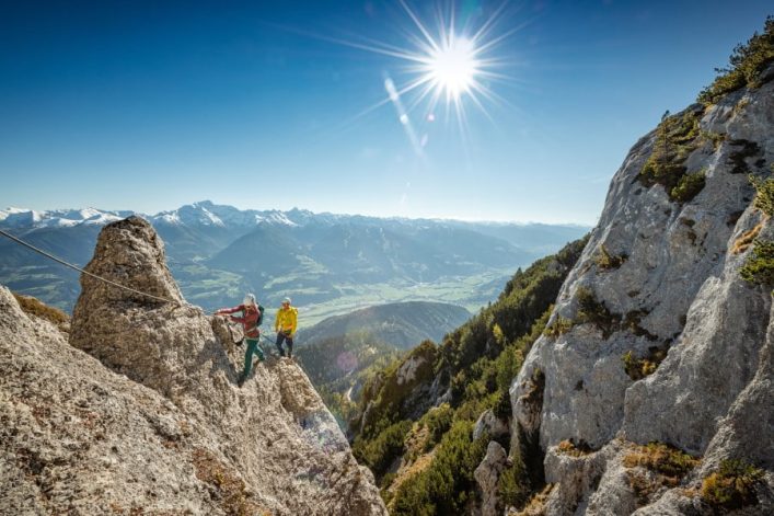 Zwei Kletterer erklimmen einen felsigen Steilhang in den Alpen bei strahlendem Sonnenschein. Der linke trägt rote Kleidung und der rechte gelbe. Im Hintergrund sind grüne Wälder und schneebedeckte Berge sichtbar.