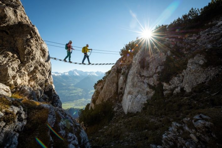 Zwei Bergsteiger überqueren eine Drahtseilbrücke zwischen zwei Felswänden im Sonnenlicht. Die Sonne strahlt hell neben dem Gipfel, während im Hintergrund schneebedeckte Berge und ein Tal sichtbar sind.