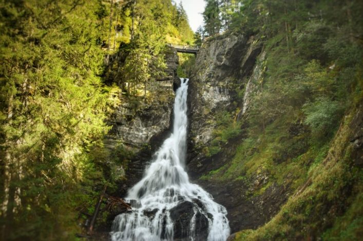 Im Zentrum fällt ein großer Wasserfall zwischen steilen Felswänden herab, umgeben von dichtem grünen Wald. Oben sieht man eine Brücke, die den Wildbach überquert.