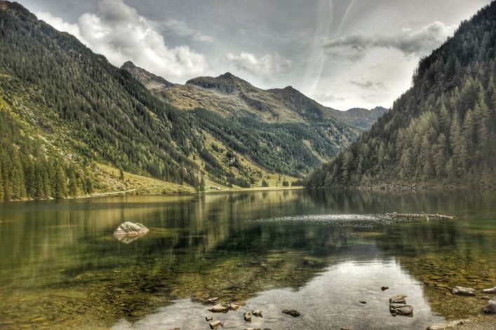 Ein klarer Bergsee in der Mitte, umgeben von bewaldeten Bergen unter einem teils bewölkten Himmel. Im Vordergrund einige Steine und Felsen am Ufer, die durch das ruhige Wasser gespiegelt werden.