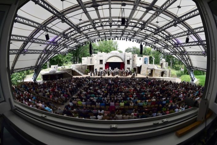 Blick von hinter dem Zuschauerraum auf die Freilichtbühne der Fränkischen Passionsspiele. Ein riesiger Zeltbogen überspannt den Zuschauerraum. Auf der Bühne sind einige verkleidete Dorfbewohner zu sehen.