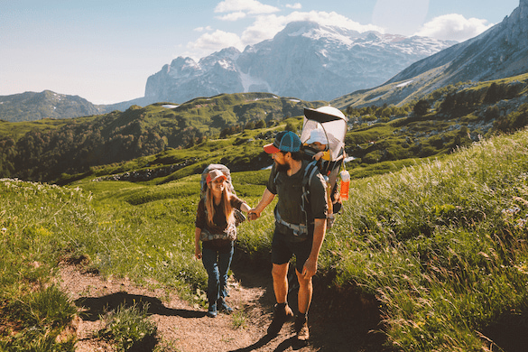 Familienspaziergang in den Bergen: Ein Vater trägt ein Kind in einer Kraxe, während er Händchen haltend mit seiner Tochter wandert. Ideal für Elternzeit Reisen, die Natur im Hintergrund ist beeindruckend.