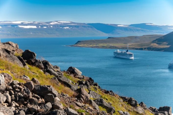 Foto eines Kreuzfahrtschiffes, dass durch einen isländischen Fjord fährt, vorbei an weiten Flächen und in Richtung hoher Berge.