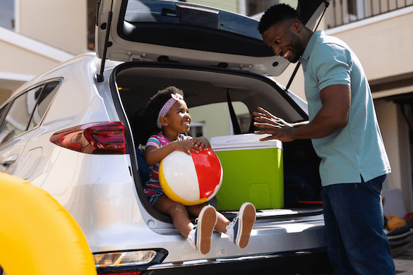 Ein Mann und seine Tochter packen den Kofferraum für den Familienurlaub. Die Tochter sitzt im geöffneten Kofferraum des Autos und hält einen Ball. Der Vater lächelt sie an und steht daneben. Elternzeit Reisen.