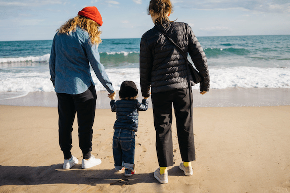 Am Strand stehen zwei Frauen mit einem Kleinkind, das ihre Hände hält, und blicken aufs Meer. Sie genießen die Sonne und das Wasser, vermutlich auf einer Elternzeit-Reise.