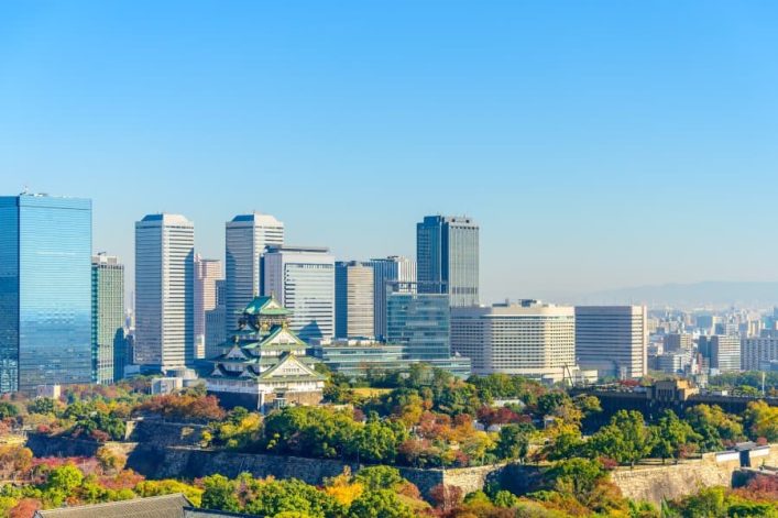 Osakas Skyline mit modernen Hochhäusern erhebt sich hinter der historischen Osaka-Burg, die inmitten grüner Bäume und Herbstfarben der Parklandschaft liegt, unter einem klaren blauen Himmel.