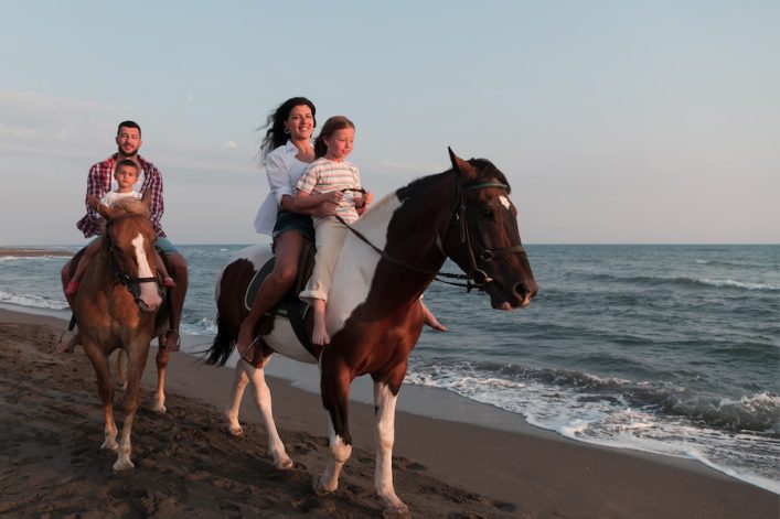 Familienurlaub auf Mallorca: Zwei Erwachsene und zwei Kinder reiten auf Pferden am sandigen Strand entlang, der Ozean im Hintergrund, während die Sonne sanft scheint. Alle wirken glücklich und entspannt.