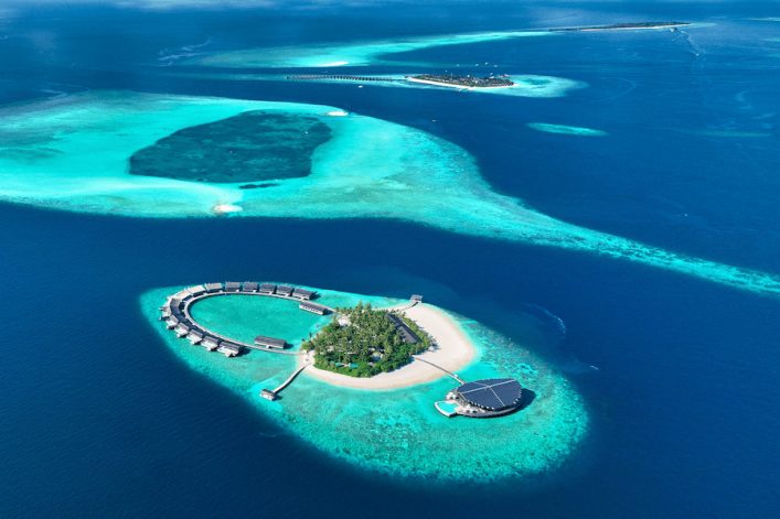 Luftaufnahme einer maledivischen Inselgruppe mit türkisfarbenem Wasser und weißen Sandstränden. Die vordere Insel ist kreisförmig bebaut. Ideal für einen Familienurlaub auf den Malediven.