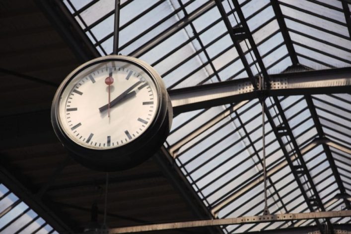 Eine große runde Uhr hängt im Bahnhof unter einem gläsernen, stahlverstärkten Dach. Die Uhr zeigt kurz nach elf Uhr und hat einen roten Sekundenzeiger. Das Dach besteht aus vielen parallelen Streben.