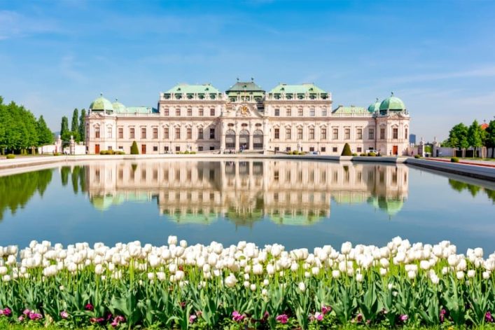 Das Belvedere in Wien im Frühling, mit weißen Tulpen im Vordergrund, einem ruhigen Spiegelteich und dem prächtigen Schloss im Hintergrund. Der klare Himmel lädt zu einer April-Kreuzfahrt ein.
