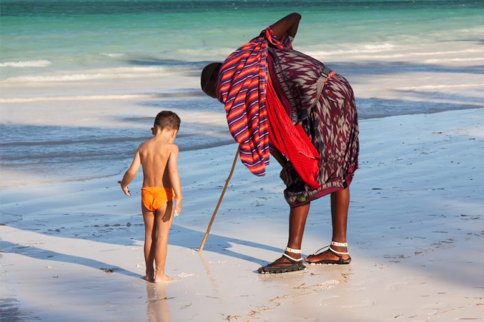 Ein Massai mit buntem Gewand und einem Stock zeigt einem kleinen Jungen im orangefarbenen Badeanzug etwas im Sand am Strand von Sansibar. Ideal für einen Familienurlaub auf Sansibar.