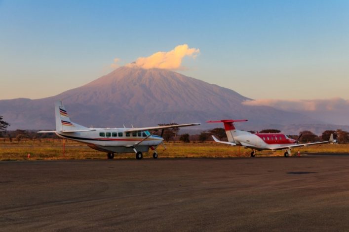 Kleine Flugzeuge stehen auf einem Rollfeld bei Sonnenuntergang. Im Hintergrund erhebt sich majestätisch ein imposanter Berg. Vorne ist Grasland zu sehen, der Himmel leuchtet sanft.