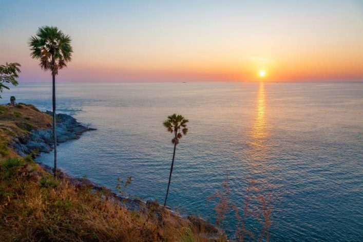 Sonnenuntergang über dem Meer an Kap Laem Promthep, Phuket. Im Vordergrund mehrere Palmen auf einer Klippe, das ruhige Wasser erstreckt sich bis zum Horizont, wo die Sonne untergeht.