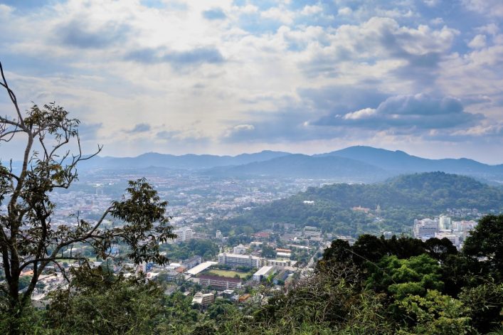 Blick vom Monkey Hill in Phuket: Im Vordergrund sind Bäume, darunter eine Stadt mit Gebäuden. Im Hintergrund erheben sich sanfte, bewaldete Hügel unter einem bewölkten Himmel.