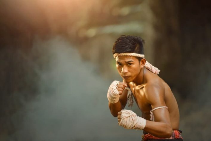 Muay Thai Boxer in angespannter Kampfpose, trägt traditionelles Stirnband und Handbandagen, Hintergrund verschwommen, warmes Licht von links oben. Dynamische und konzentrierte Körperhaltung.