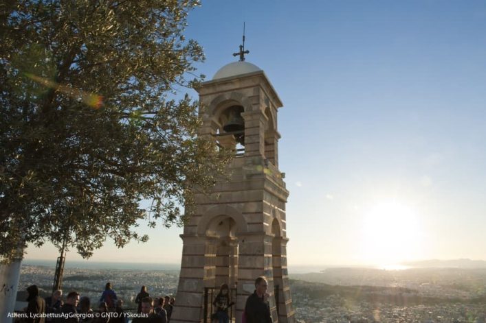 Turm mit Kreuz unter blauem Himmel; Sonnenuntergang im Hintergrund. Menschen stehen rund um den Turm, der von einem Baum teilweise verdeckt wird. Blick auf eine weite Stadtlandschaft.
