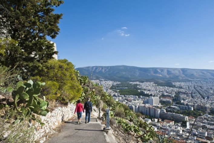 Zwei Personen spazieren auf einem Pfad auf dem Lycabettus Hügel, mit Blick auf Athen im Hintergrund. Links wachsen Bäume und Kakteen, rechts erstreckt sich die Stadt unter einem klaren Himmel.
