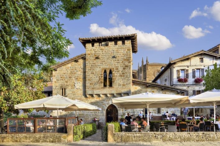 Historisches Steingebäude mit Arkadenfenstern in der Mitte, von Sonnenschirmen und einem Café im Freien umgeben. Blauer Himmel mit weißen Wolken im Hintergrund, links Baumäste und rechts blühende Pflanzen.