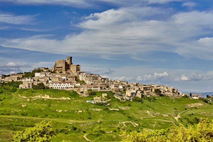 Auf einem grünen Hügel liegt das malerische Dorf Ujué in Navarra, Spanien. Oben dominiert eine beeindruckende Kirche, umgeben von dicht gedrängten Steinhäusern unter einem bewölkten Himmel.