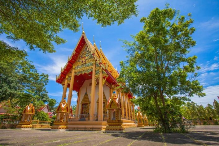 Ein buddhistischer Tempel in lebendigen Farben steht in der Mitte, umgeben von Bäumen unter klarem, blauem Himmel. Der Boden ist betoniert und die Architektur ist reich verziert.