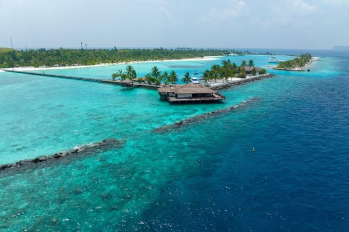 Tropische Insel mit Palmen am türkisfarbenen Meer, ein perfektes Ziel für Flitterwochen auf den Malediven. Auf Stelzen gebaute Holzhütten ragen ins Wasser, umgeben von einem langen Steg.