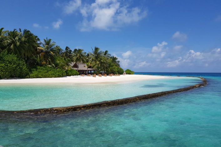 Tropischer Strand mit Palmen und klarem, türkisfarbenem Wasser. Eine kleine Sandbank ragt ins Meer. Der blaue Himmel ist mit vereinzelten Wolken bedeckt. Im Hintergrund sind Liegestühle zu sehen.