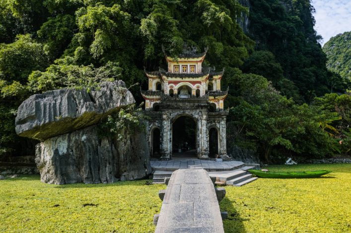 Steinpfad führt über von Entengrütze bedecktes Wasser zu einer verzierten Pagode, flankiert von üppigem Grün und imposantem Felsen. Rechts schwimmt ein grünes Kanu auf dem Wasser. Hintergrund von dichtem Wald.