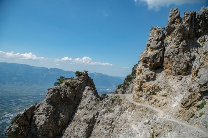 Steiniger Gebirgspfad durch raue Felslandschaft, links steiler Abgrund mit Blick ins Tal, rechts hohe Felswand. Einige Wolken am blauen Himmel.