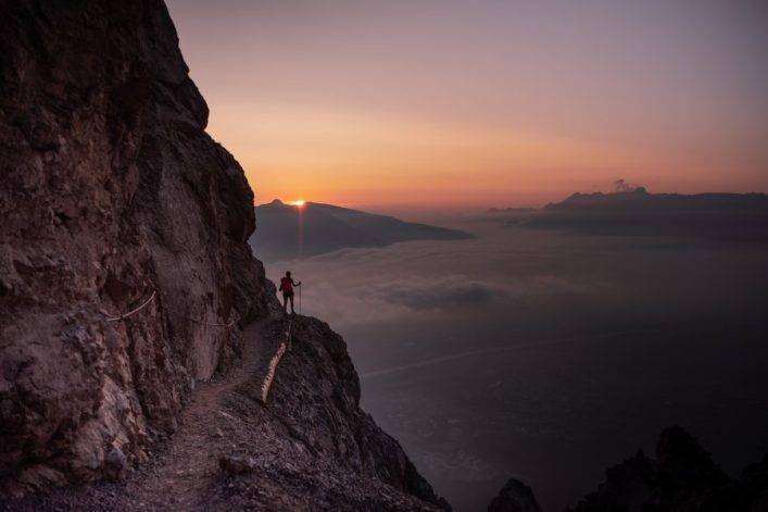 Ein schmaler Bergpfad windet sich entlang einer steilen Felswand links, während ein Wanderer im roten Outfit in der Bildmitte den Sonnenuntergang über bewölktem Tal rechts bewundert.