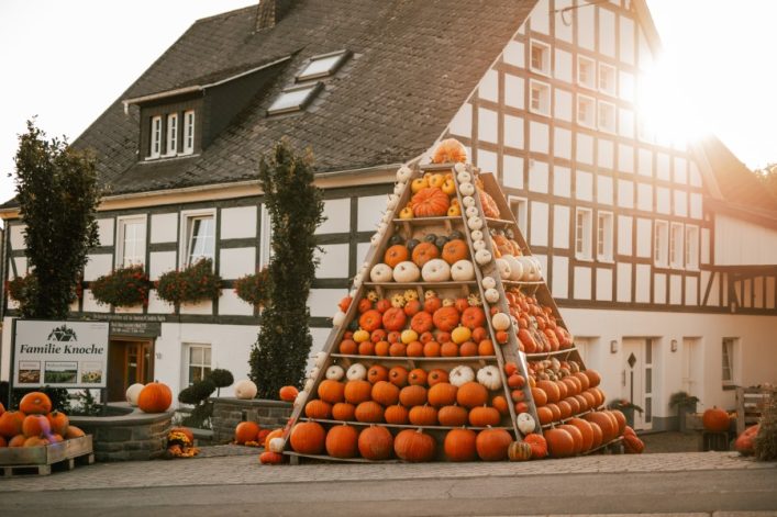 Eine Pyramide aus zahlreichen Kürbissen steht vor einem Fachwerkhaus. Rechts scheint die Sonne. Links befindet sich ein Schild mit der Aufschrift „Familie Knoche“. Blumen hängen an den Fenstern.