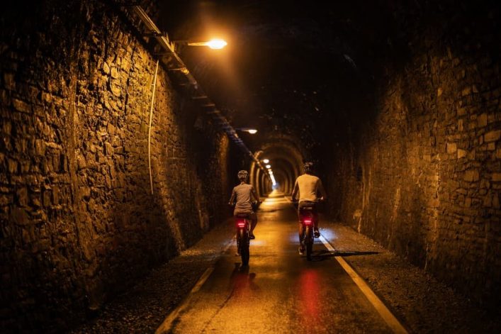 Zwei Menschen fahren mit Fahrrädern durch einen beleuchteten, mit Backsteinwänden gesäumten Tunnel. Die Beleuchtung verbreitet ein warmes Licht, das Schatten auf den Boden wirft.