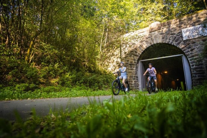 Radfahrer verlassen einen steinernen Tunnel auf einem Waldweg. Üppiges Grün umgibt die Szene, während Sonnenlicht auf das Laub scheint. Der Tunnel trägt ein Schild mit dem Text „Kückelheimer Tunnel“.